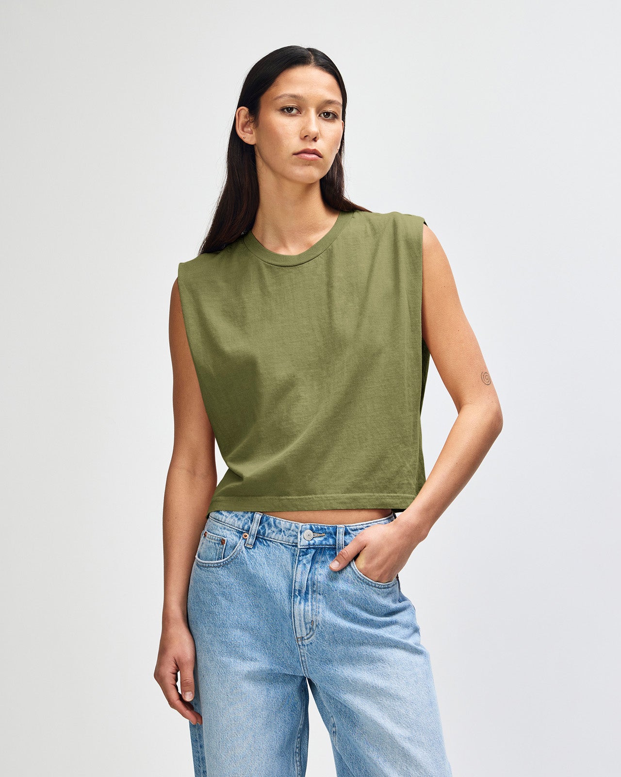 Model wearing a faded army Garment Dyed Women's Muscle Tee, sleeveless, paired with light blue jeans, standing against a plain white background (front pose) -army