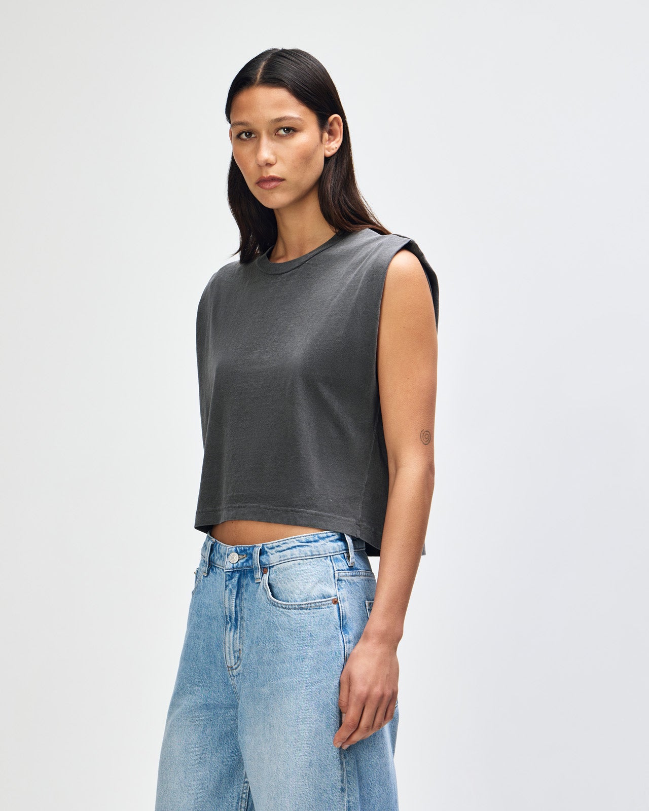 Model wearing a faded black Garment Dyed Women's Muscle Tee, sleeveless, paired with light blue jeans, standing against a plain white background (front pose) -black