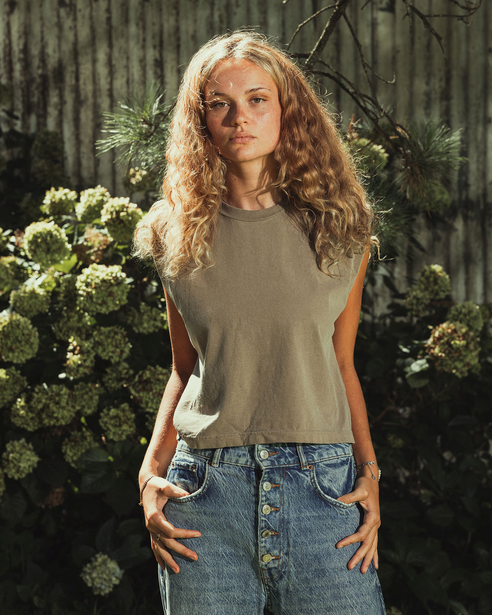 image of Women's Muscle T-Shirt : Model wearing a faded brown Garment Dyed Women's Muscle Tee, sleeveless, paired with light blue jeans, standing against a plain white background (front pose) -brown