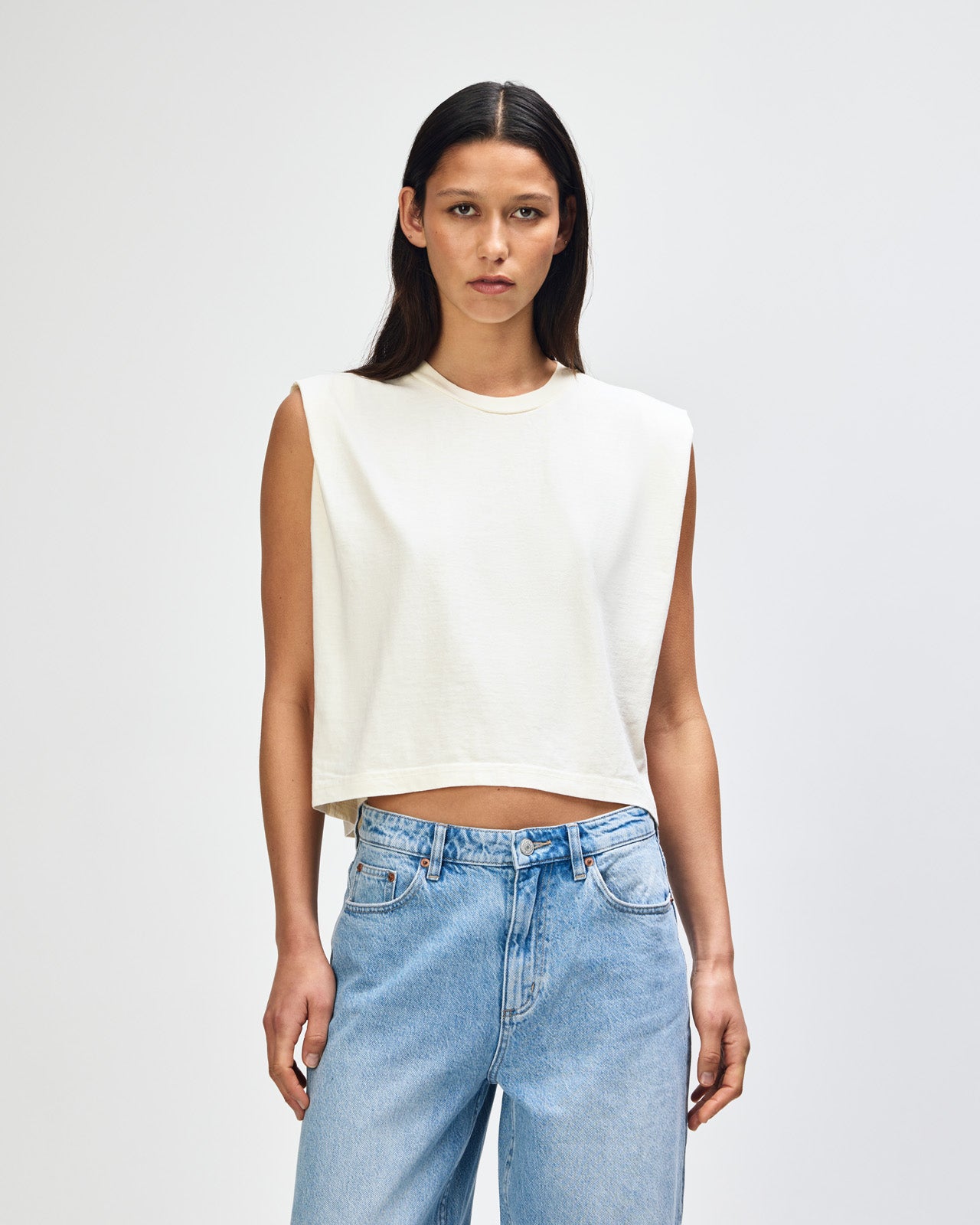 Model wearing a faded cream Garment Dyed Women's Muscle Tee, sleeveless, paired with light blue jeans, standing against a plain white background (front pose) -cream