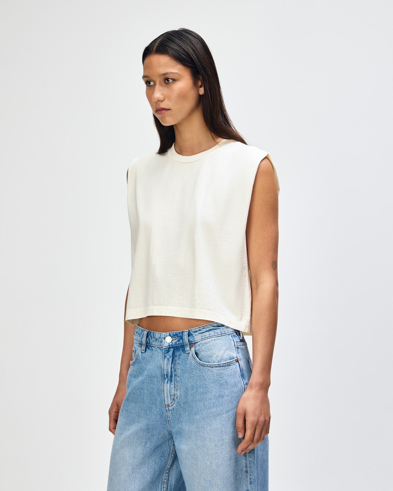 Model wearing a faded cream Garment Dyed Women's Muscle Tee, sleeveless, paired with light blue jeans, standing against a plain white background (front pose) -cream