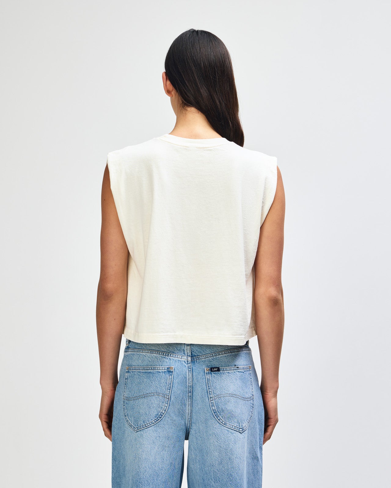 Model wearing a faded cream Garment Dyed Women's Muscle Tee, sleeveless, paired with light blue jeans, standing against a plain white background (front pose) -cream