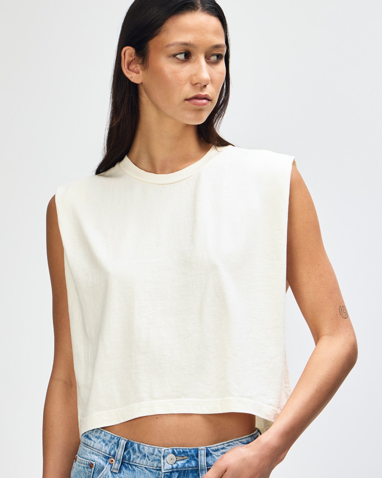 Model wearing a faded cream Garment Dyed Women's Muscle Tee, sleeveless, paired with light blue jeans, standing against a plain white background (front pose) -cream