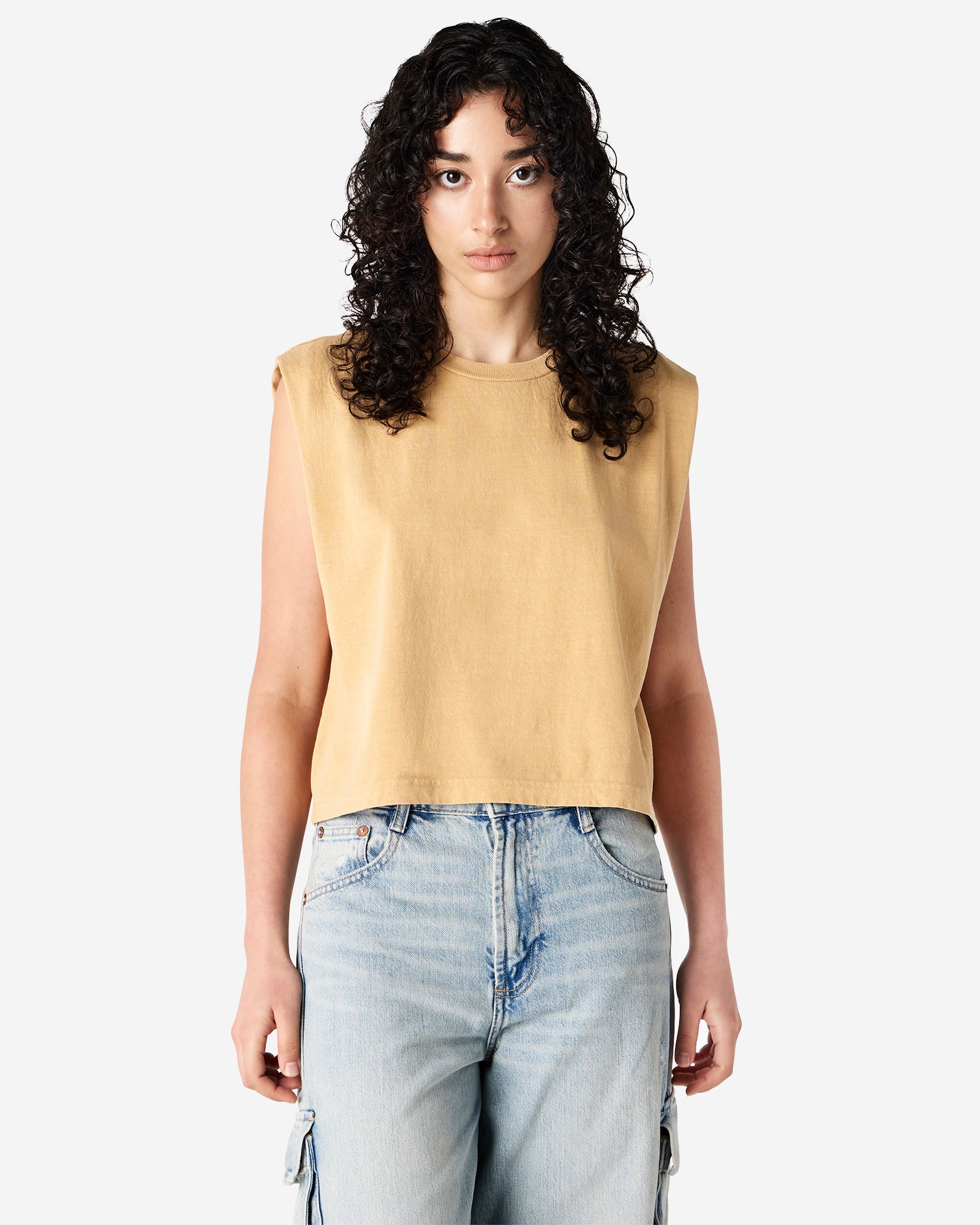 Model wearing a faded mustard Garment Dyed Women's Muscle Tee, sleeveless, paired with light blue jeans, standing against a plain white background (front pose) -mustard