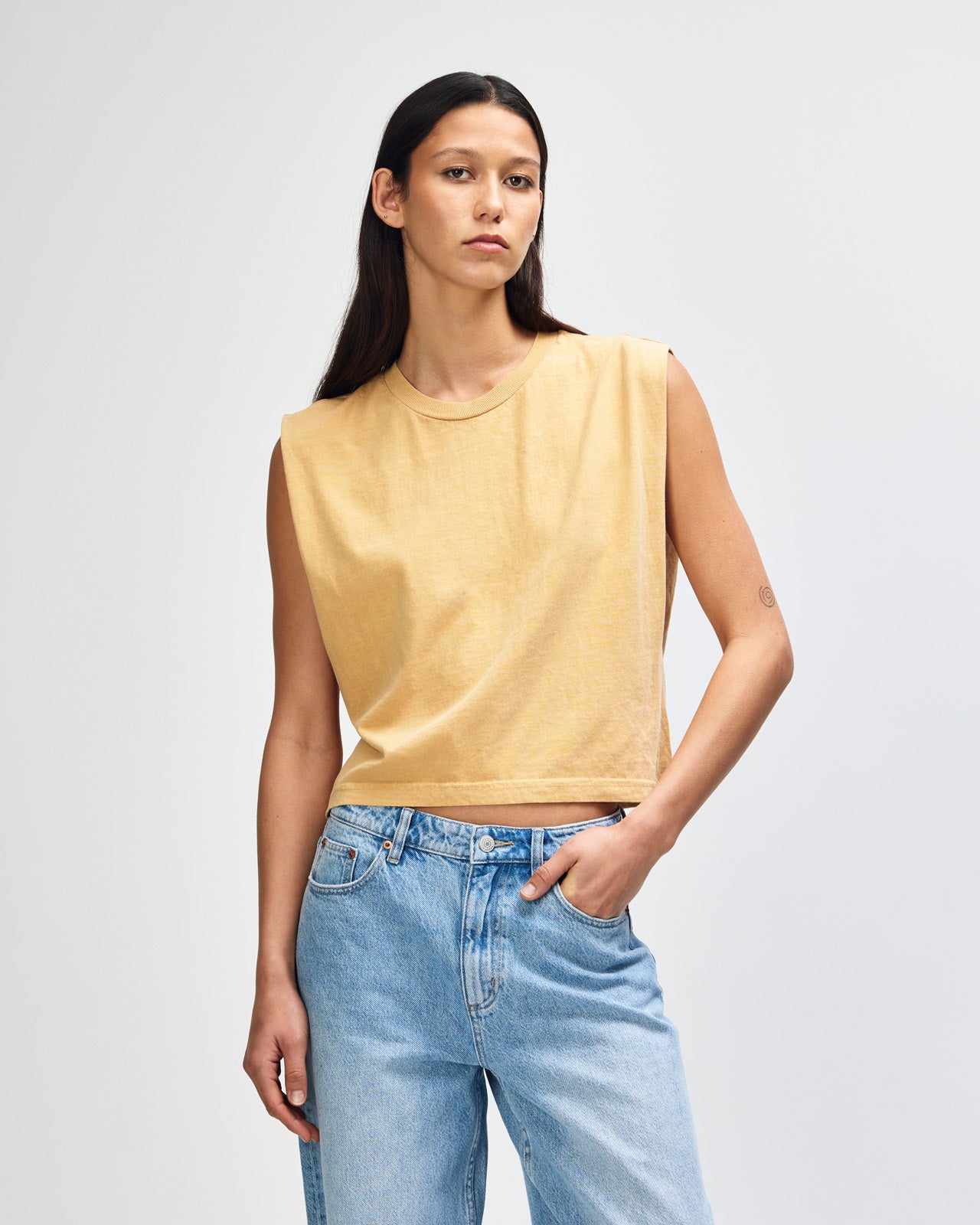 Model wearing a faded mustard Garment Dyed Women's Muscle Tee, sleeveless, paired with light blue jeans, standing against a plain white background (front pose) -mustard