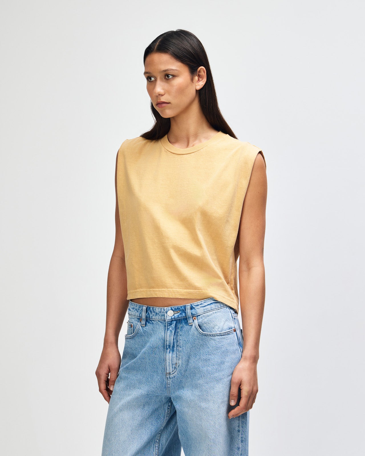 Model wearing a faded mustard Garment Dyed Women's Muscle Tee, sleeveless, paired with light blue jeans, standing against a plain white background (front pose) -mustard