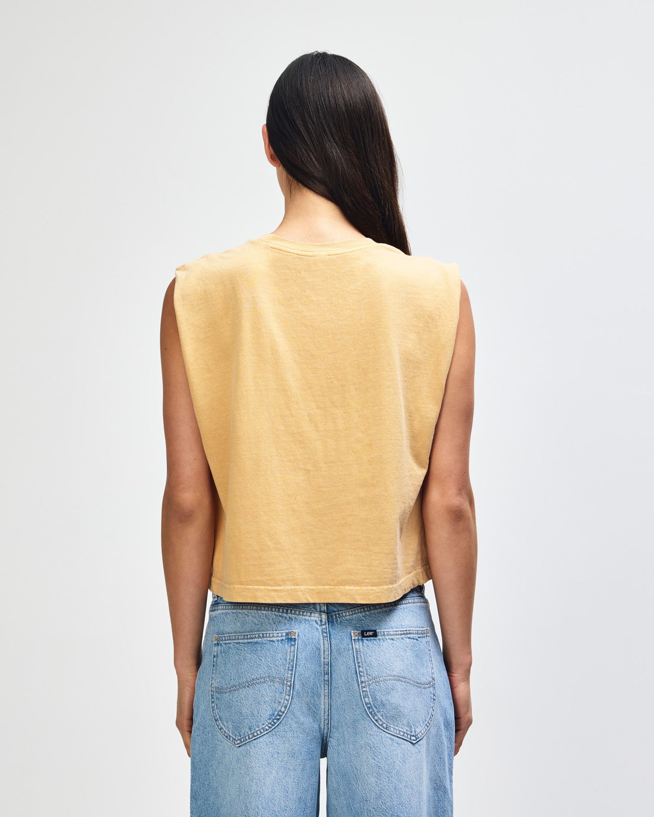 Model wearing a faded mustard Garment Dyed Women's Muscle Tee, sleeveless, paired with light blue jeans, standing against a plain white background (front pose) -mustard