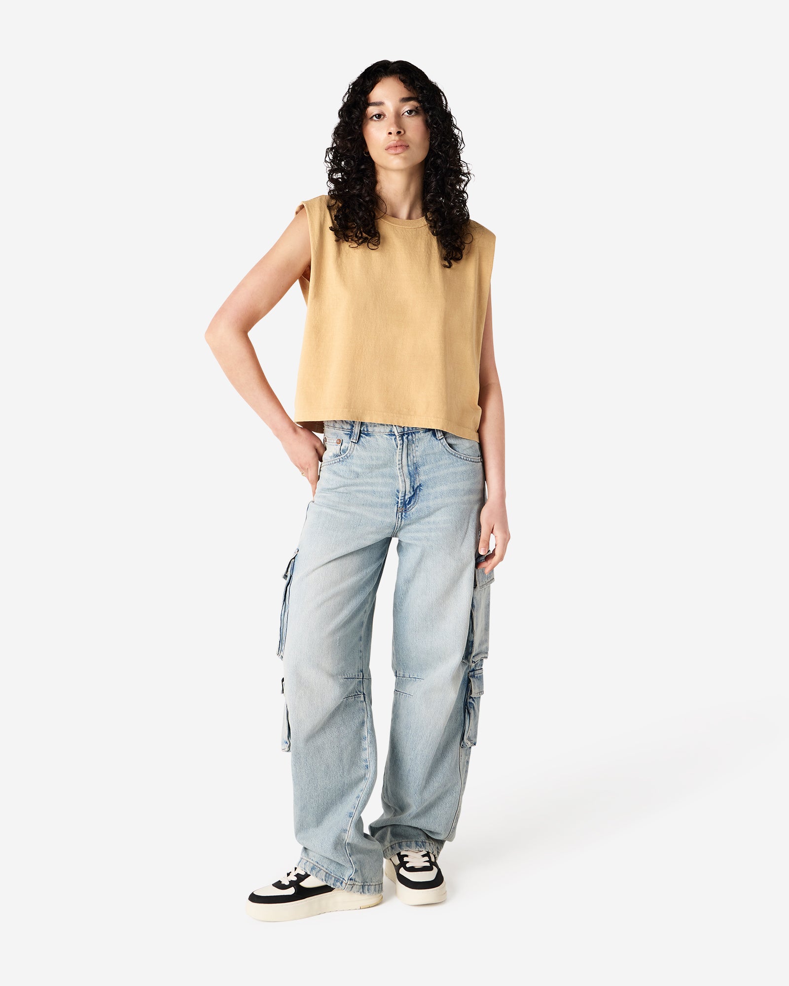 Model wearing a faded mustard Garment Dyed Women's Muscle Tee, sleeveless, paired with light blue jeans, standing against a plain white background (front pose) -mustard