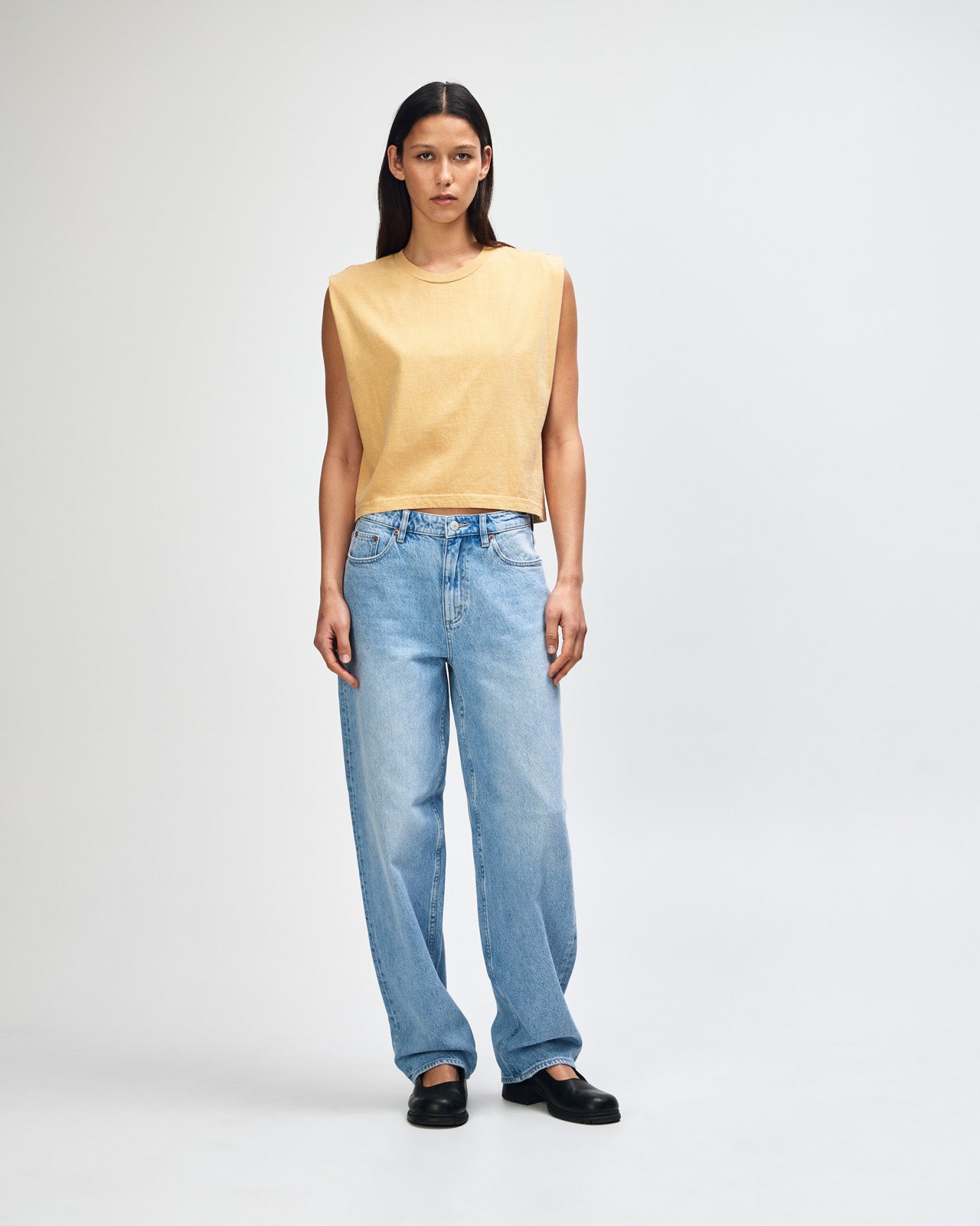 Model wearing a faded mustard Garment Dyed Women's Muscle Tee, sleeveless, paired with light blue jeans, standing against a plain white background (front pose) -mustard