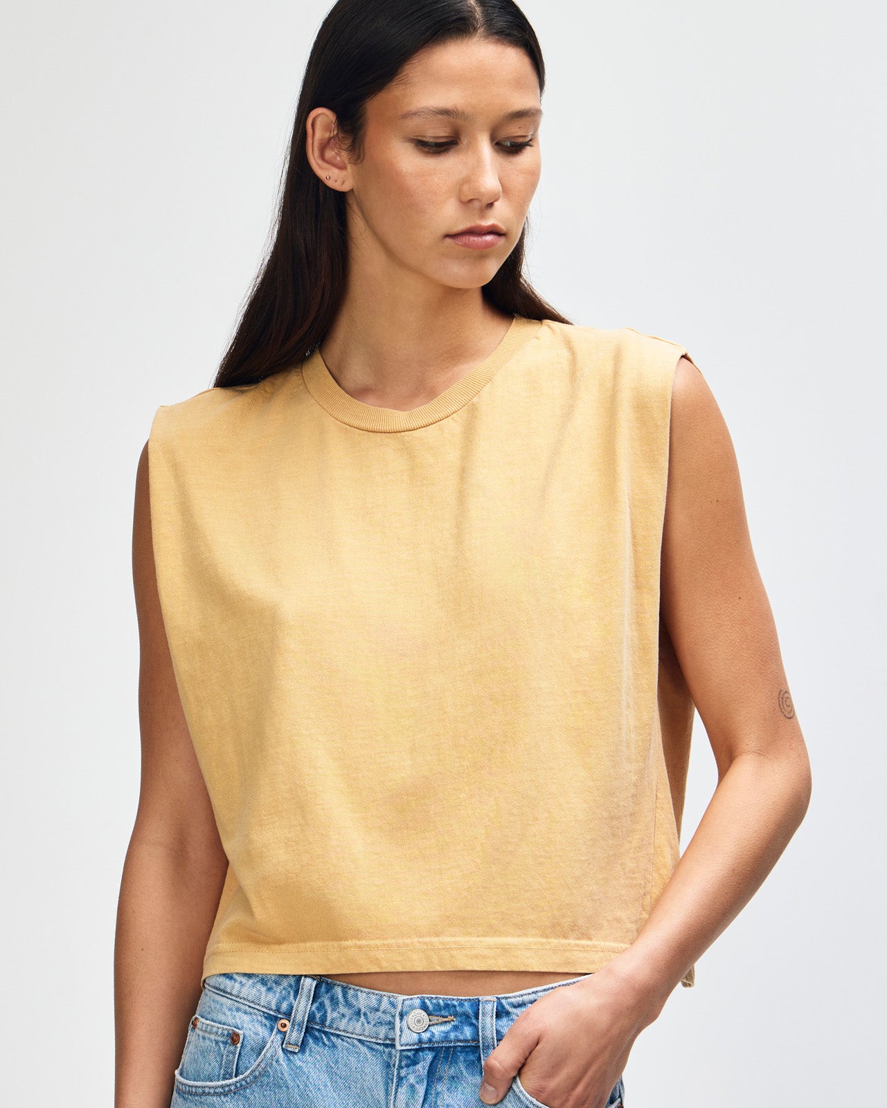 Model wearing a faded mustard Garment Dyed Women's Muscle Tee, sleeveless, paired with light blue jeans, standing against a plain white background (front pose) -mustard