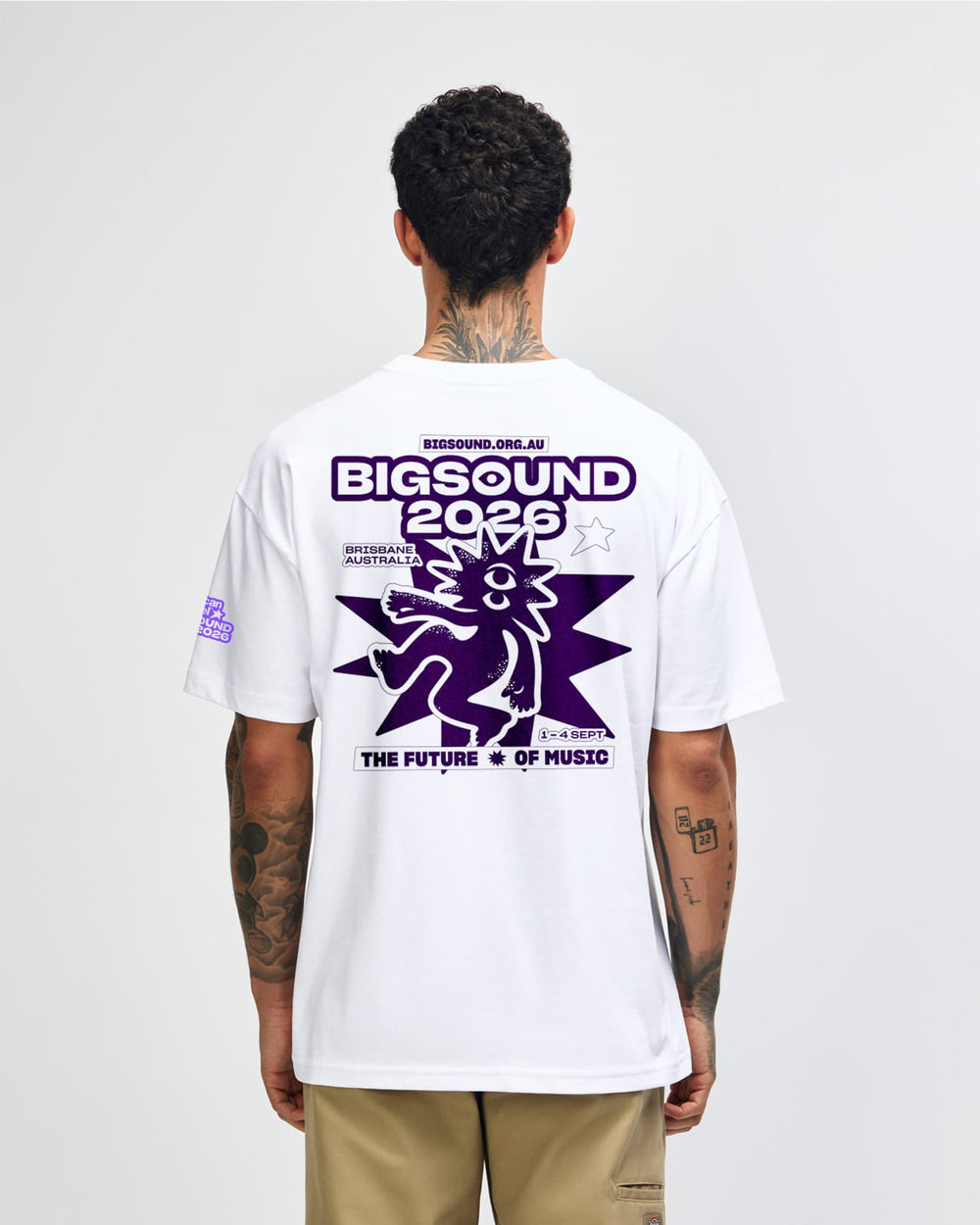 next image of BIGSOUND Lineup T-Shirt - White : A man viewed from behind wearing a white t shirt with a large purple graphic print on the back that reads BIGSOUND 2026 and THE FUTURE IS OF MUSIC -white