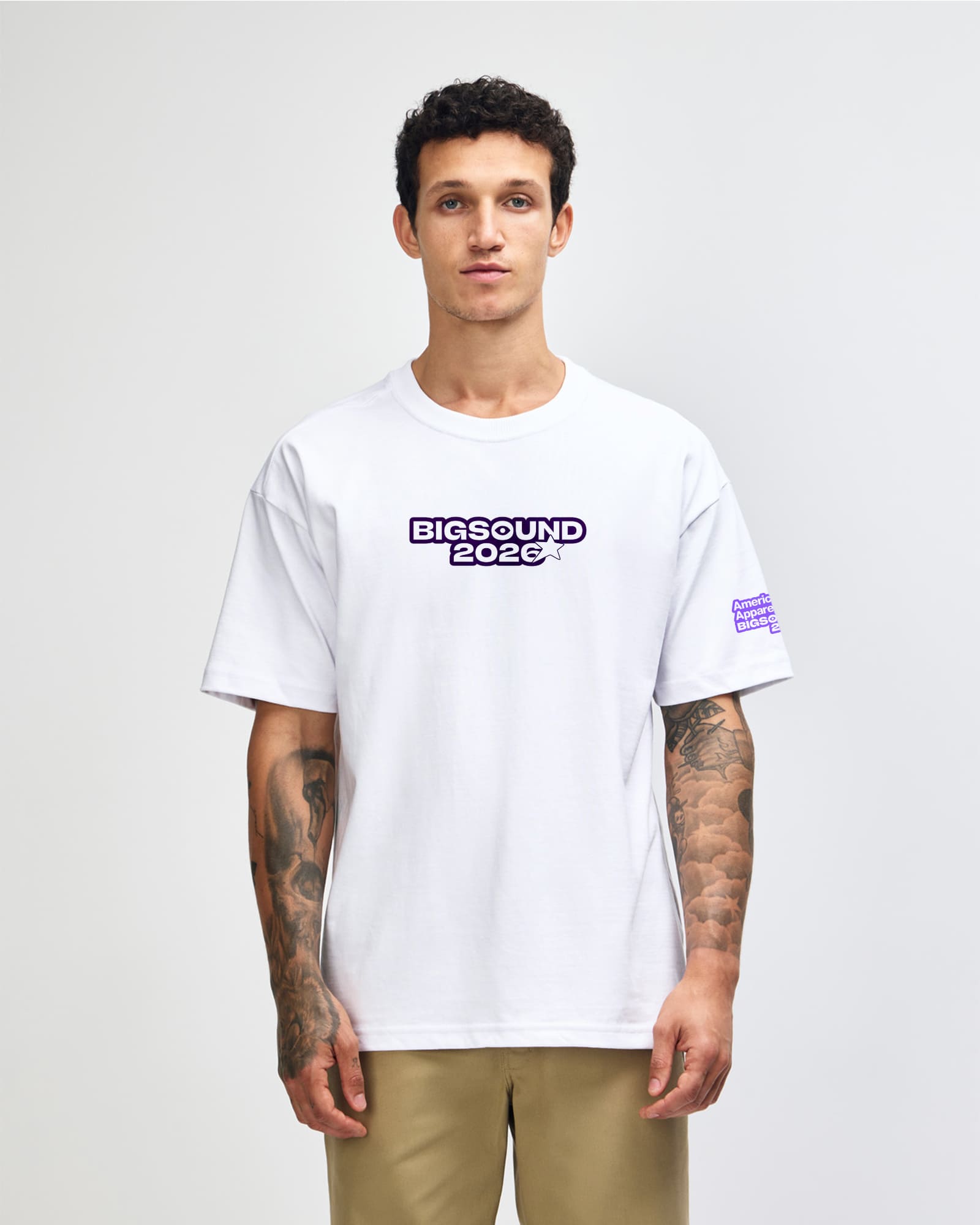 A man wearing a white oversized t shirt with BIGSOUND 2020 printed in purple on the chest, paired with khaki pants against a light gray studio background -white
