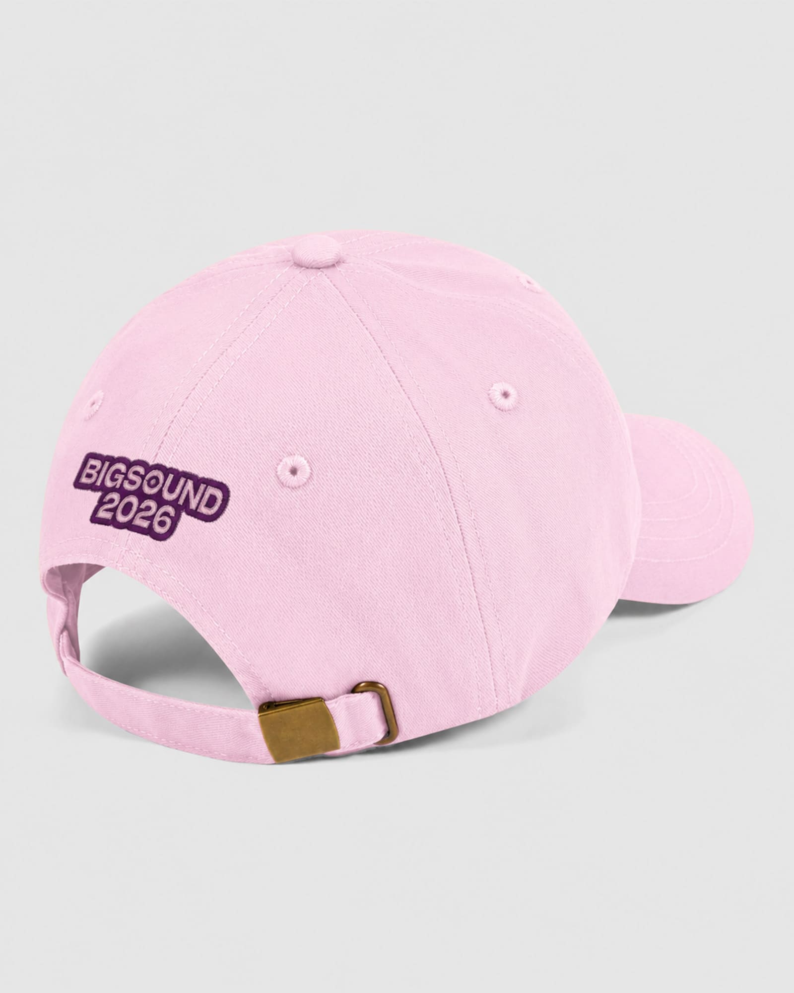 A light pink baseball cap shown from a three quarters back angle on a light gray background. The cap features 'BIGSOUND 2026' embroidered in white text inside a dark purple oval patch on the side, with a matching pink adjustable strap and a small gold metal buckle -pink