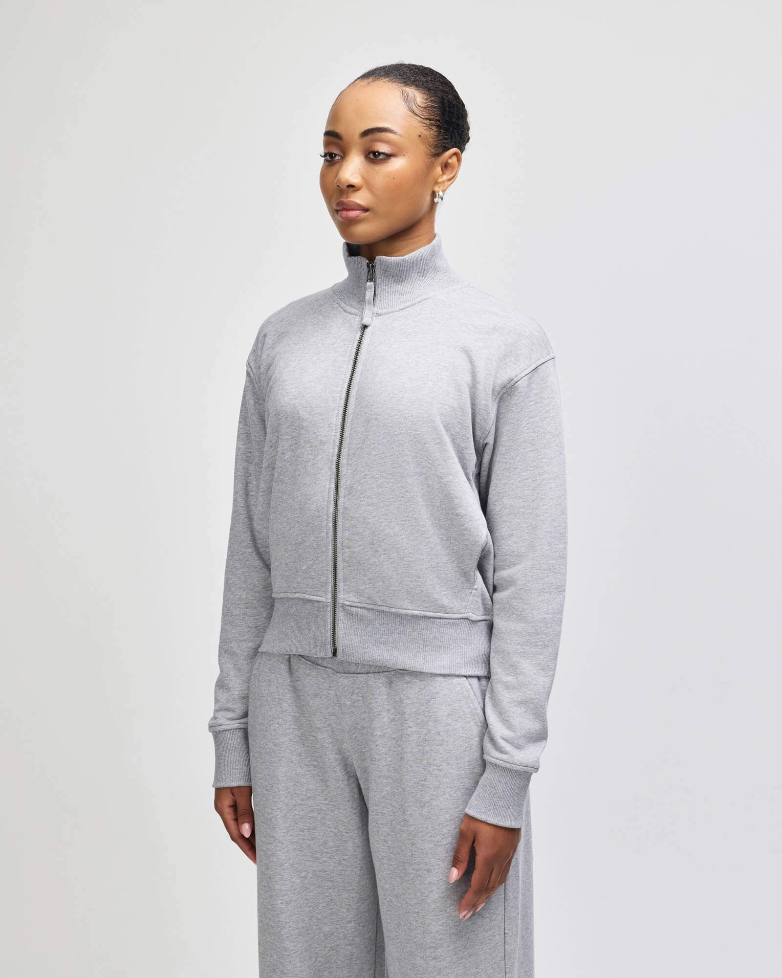 A woman modeling a light gray heather zipup sweatshirt and matching sweatpants against a plain light gray studio background -heather grey
