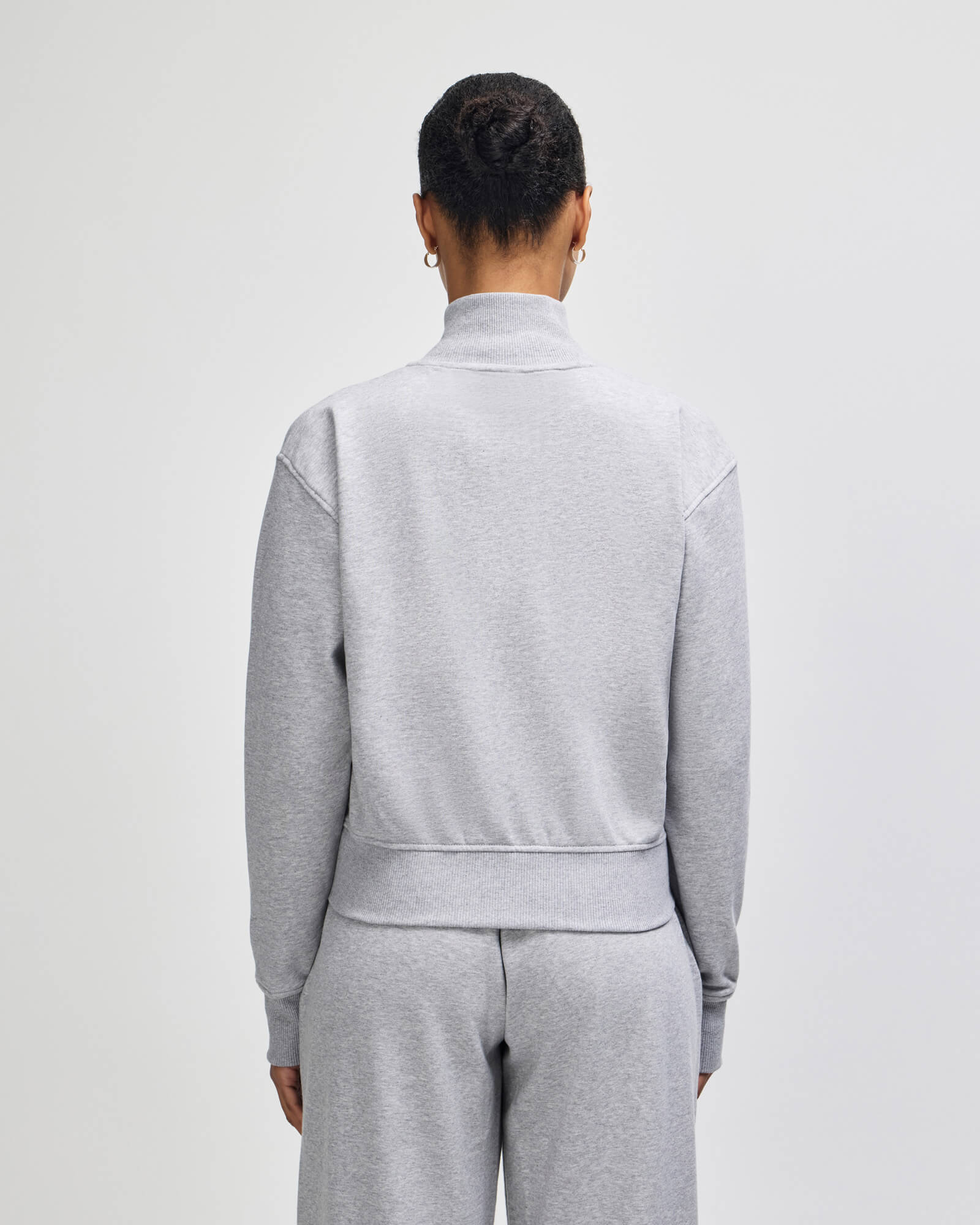 A person shown from the back wearing a light gray heathered mockneck sweatshirt and matching sweatpants against a plain light gray studio background -heather grey