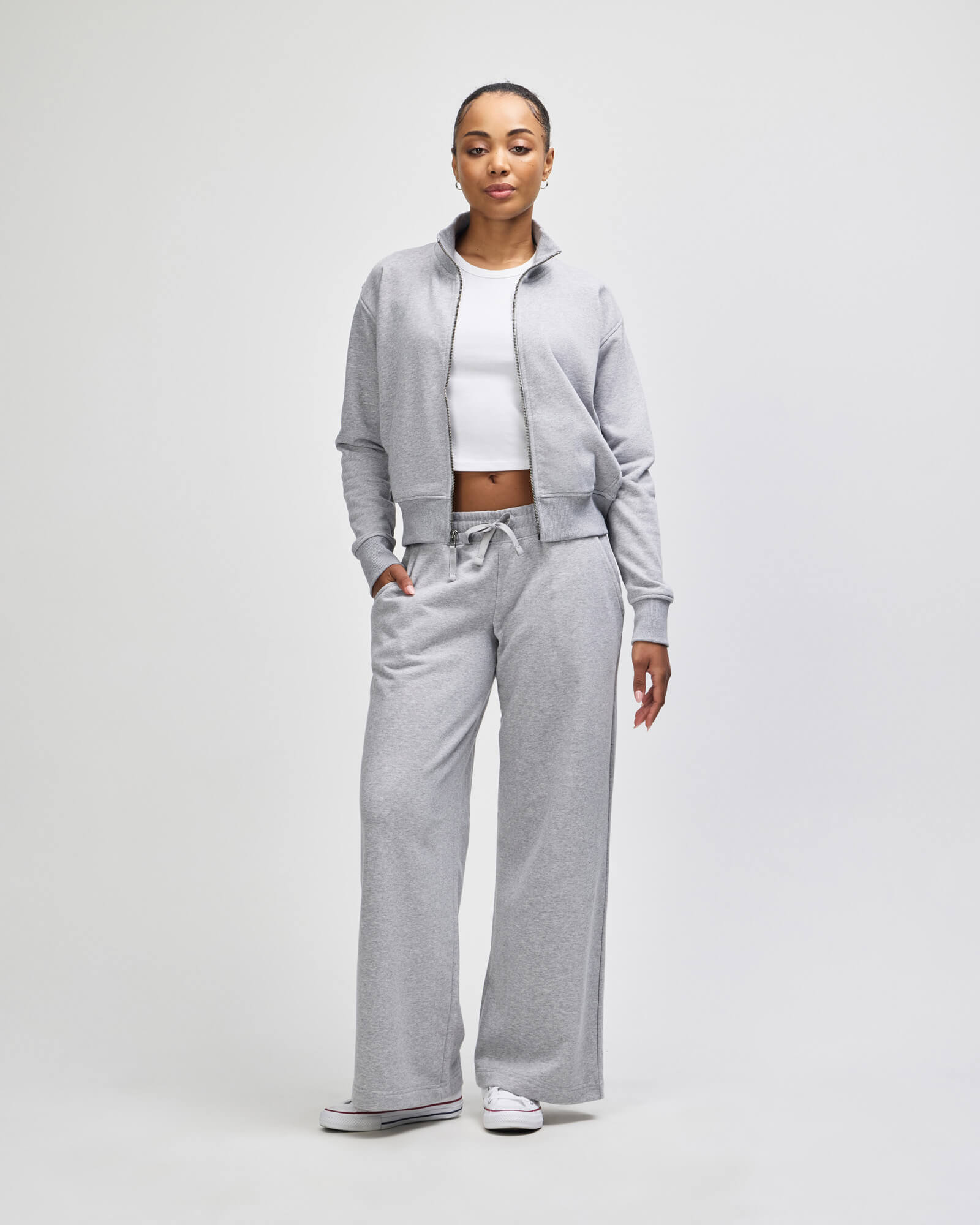 A woman modeling a light gray twopiece tracksuit consisting of a zipup hoodie and matching wideleg sweatpants, styled with a white crop top and white sneakers against a light gray studio background -heather grey