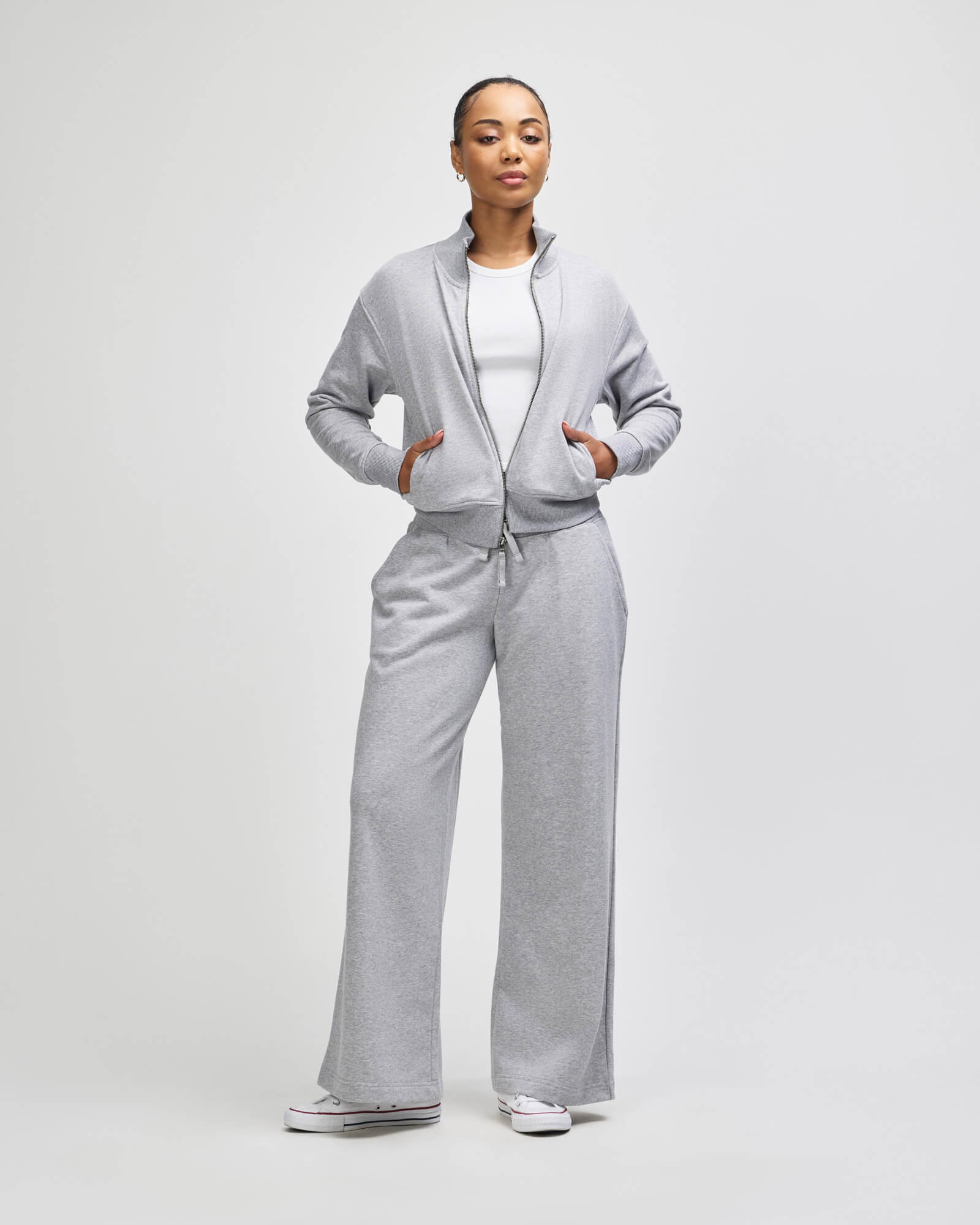 A woman modeling a light gray zipup hoodie and matching wideleg sweatpants, styled over a white top and white sneakers, against a plain light gray studio background -heather grey