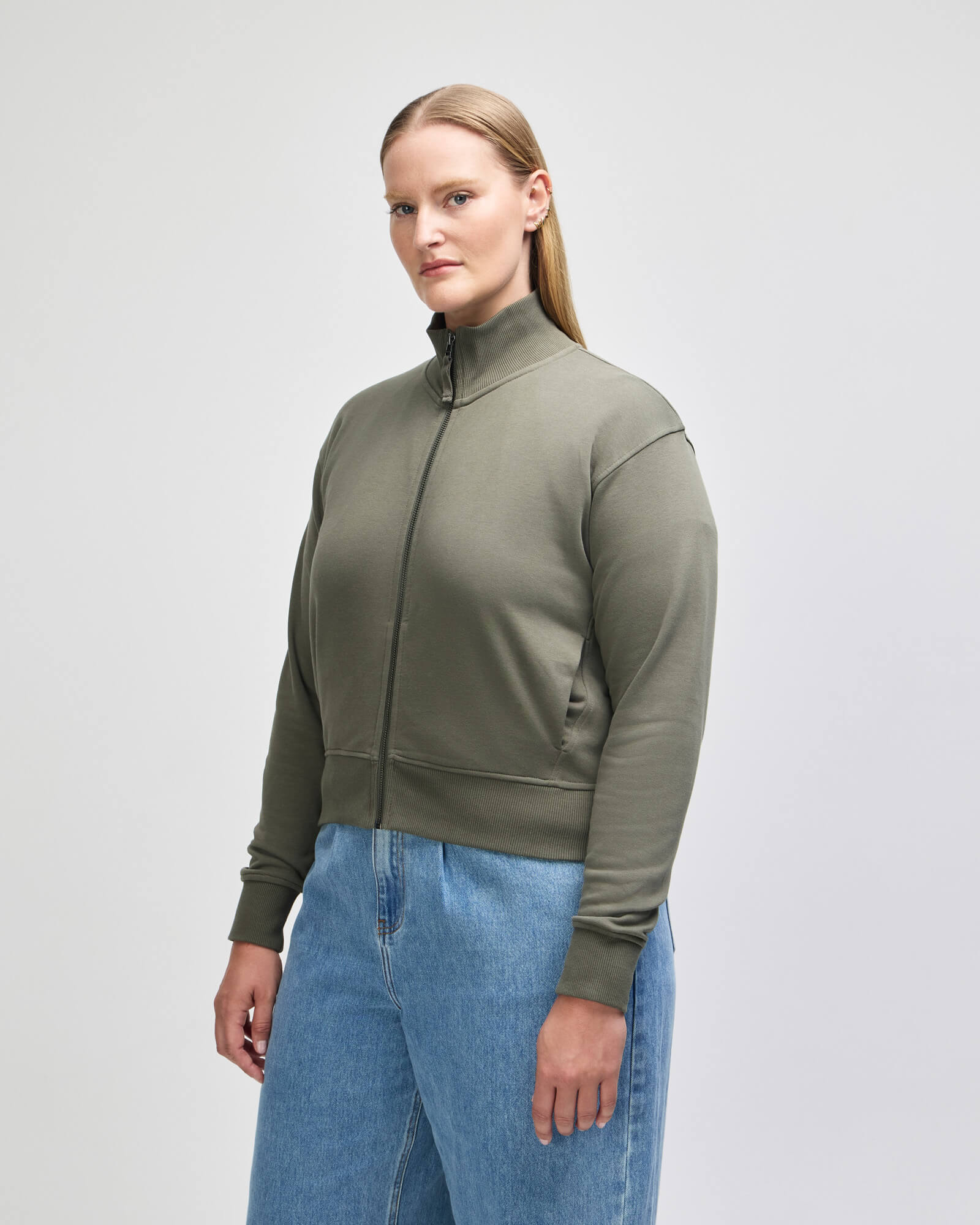 A woman modeling a cropped, olive green zipup jacket with a mock neck and ribbed cuffs, paired with light blue jeans, against a plain light gray studio background -lieutenant