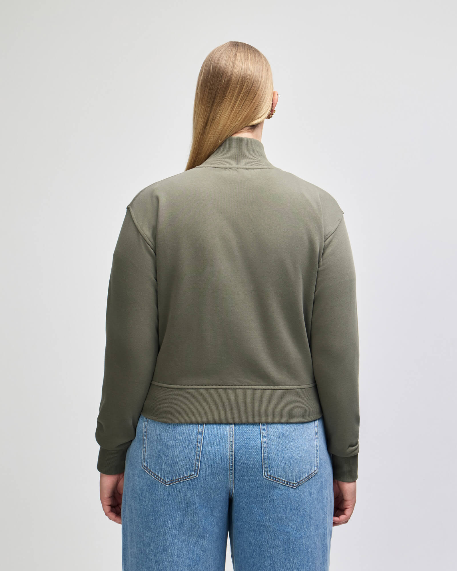 A woman viewed from the back wearing an olive green mockneck longsleeve top and light blue jeans against a light gray studio background.-lieutenant