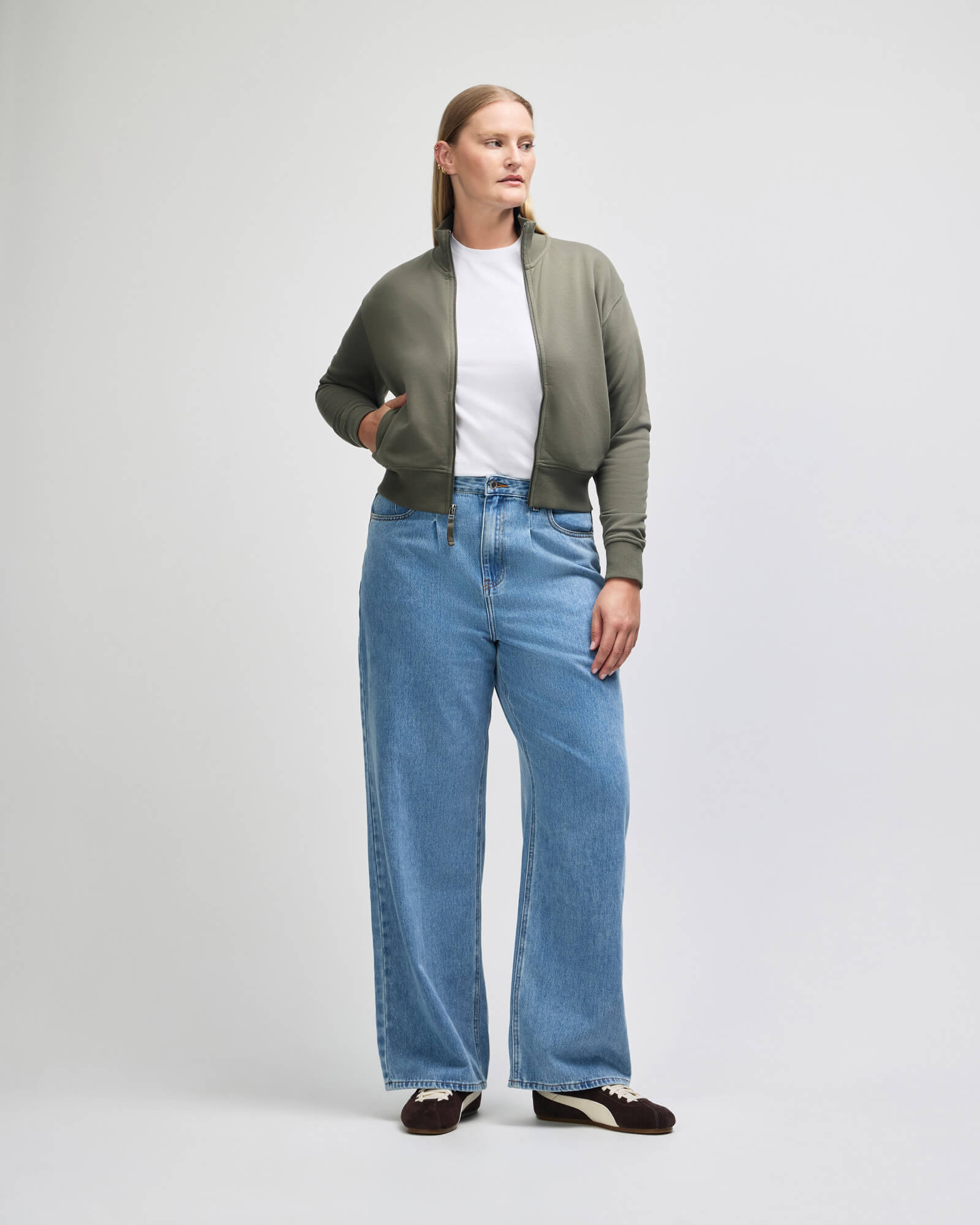 A woman modeling a light olive green bomber jacket over a white tshirt and highwaisted, wideleg blue jeans, standing against a neutral light gray studio background -lieutenant