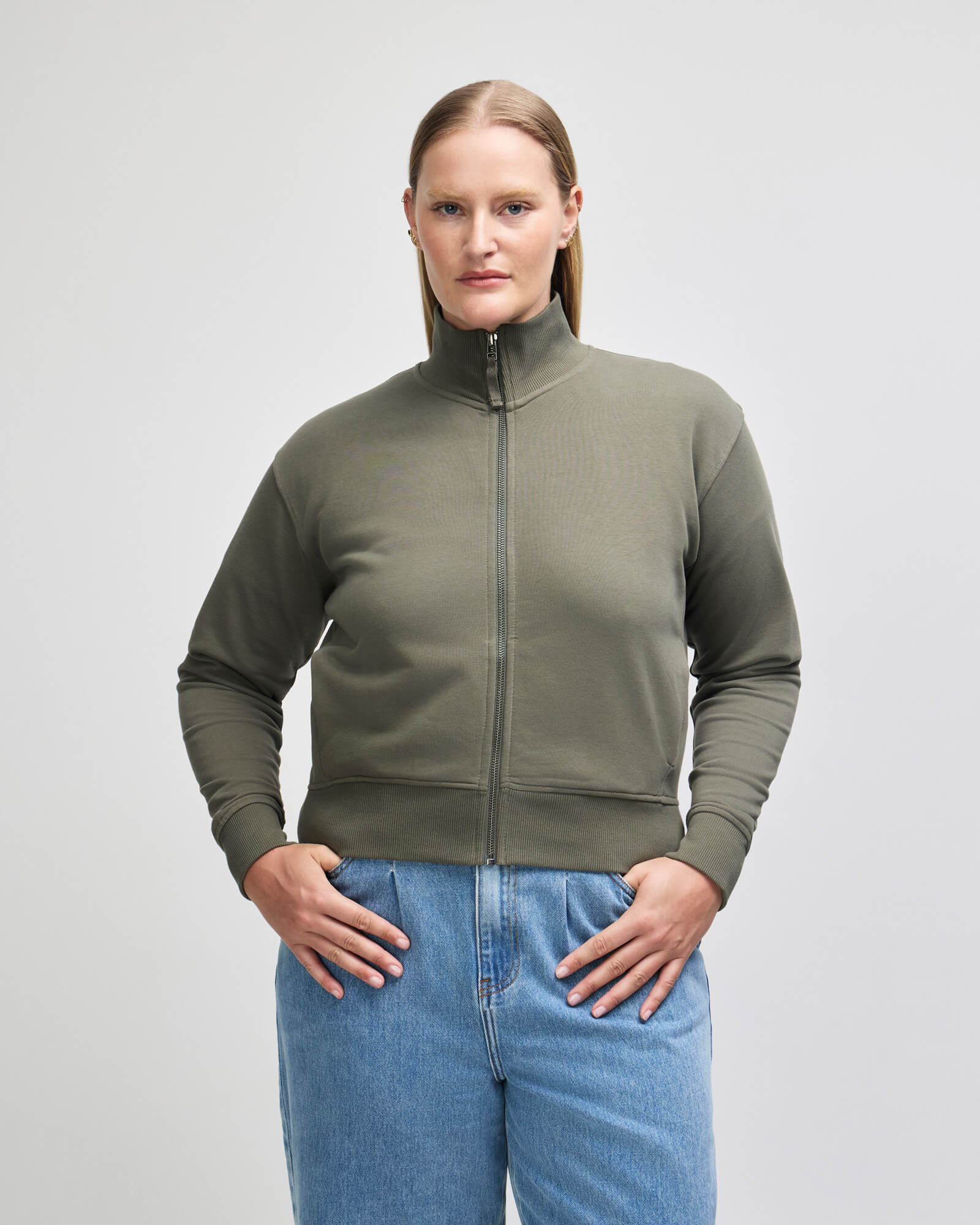 A woman modeling a cropped, olive green zipup jacket with a mock neck and ribbed cuffs, paired with light blue jeans, against a plain light gray studio background -lieutenant