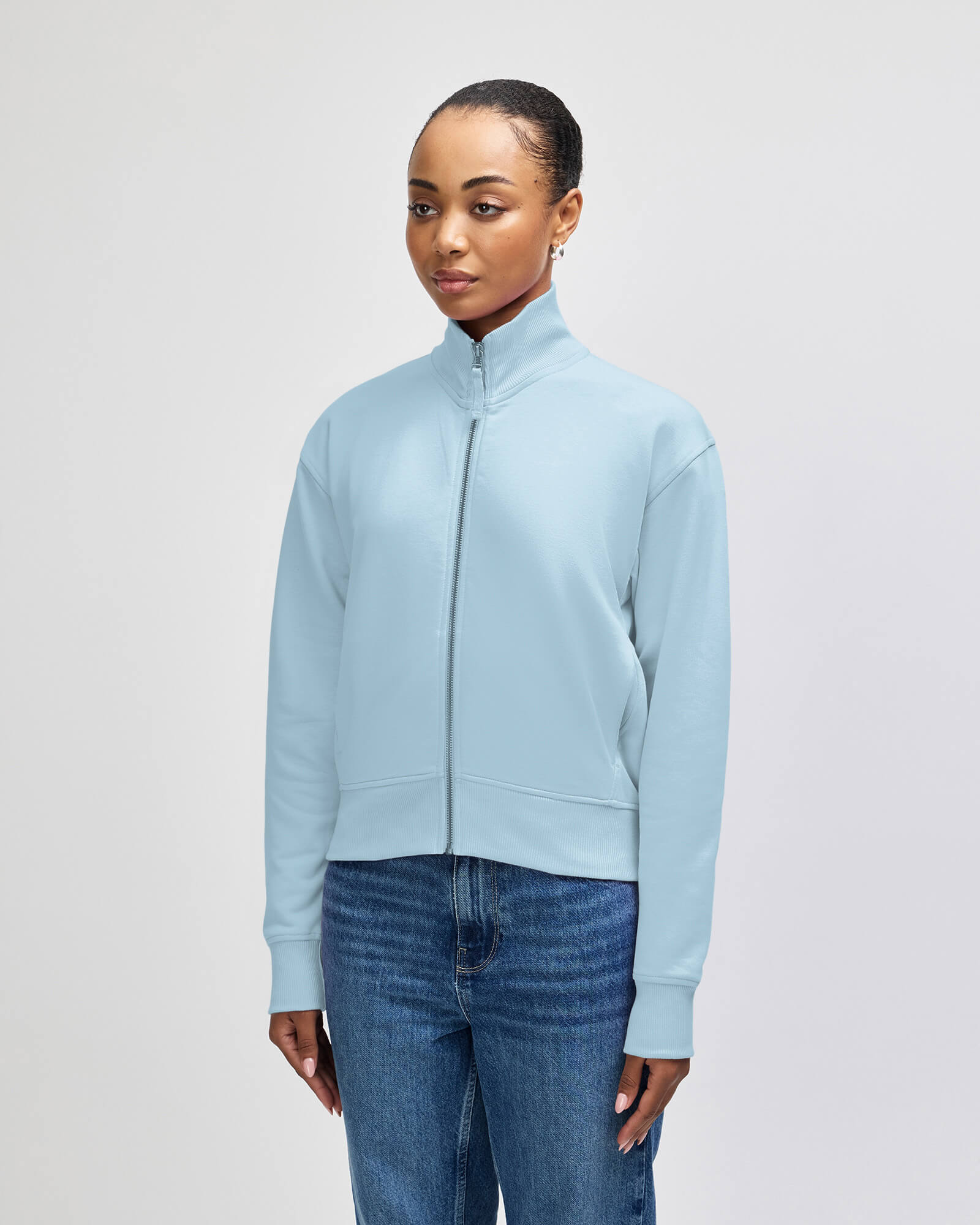 A woman modeling a light blue zipup mock neck jacket with ribbed cuffs and hem, paired with blue jeans, against a plain light gray studio background -powder blue