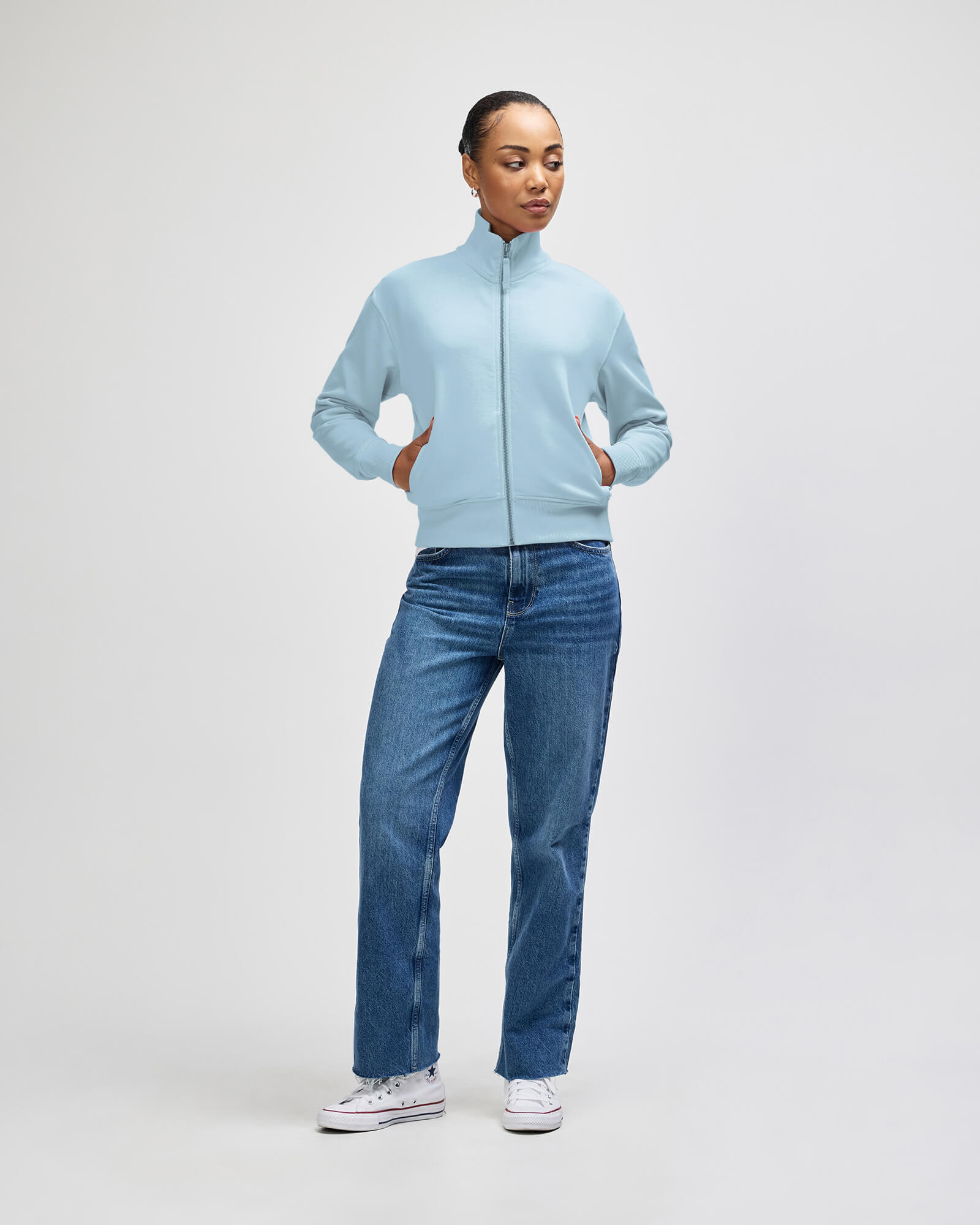 A woman modeling a light blue zipup jacket with a mock neck, paired with blue jeans and white sneakers, against a plain light gray studio background -powder blue