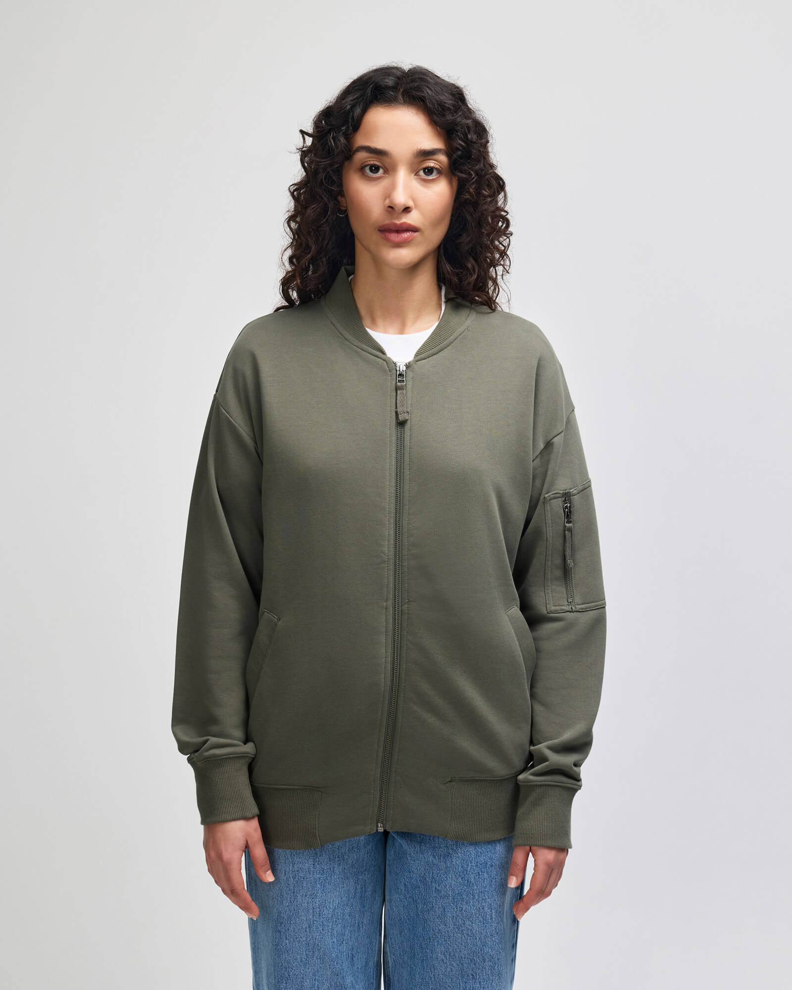 A woman with curly dark hair wearing an olive green zip-up bomber jacket over a white t-shirt and blue jeans, standing against a light gray studio background.-lieutenant