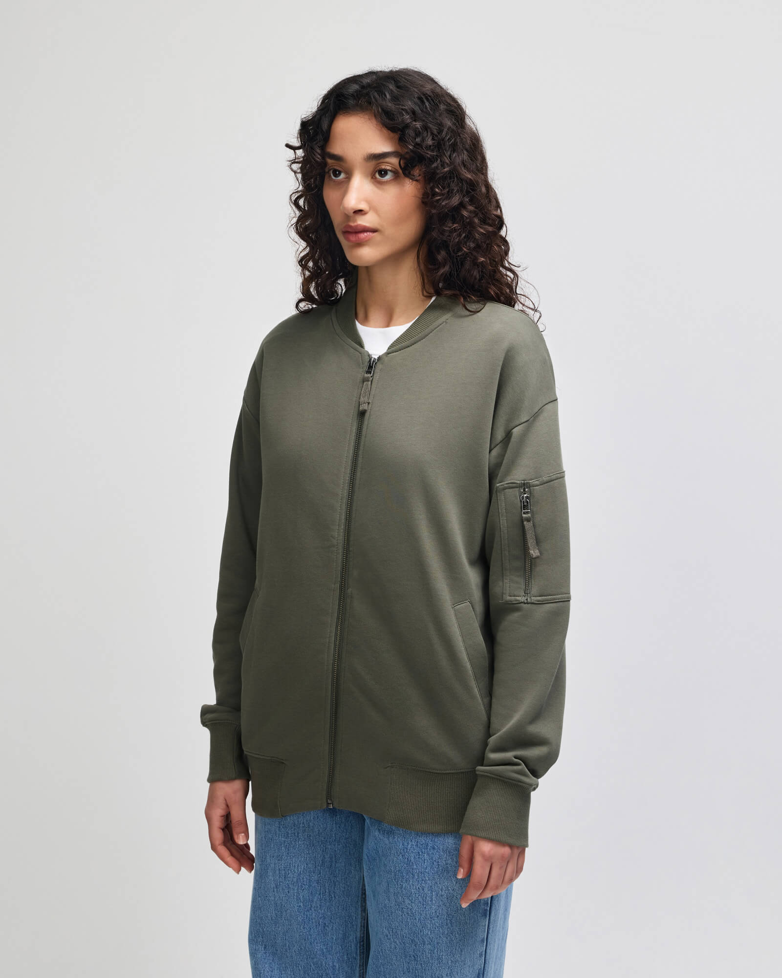 A woman with curly dark hair wearing an olive green zipup hoodie with a stand-up collar, ribbed cuffs and hem, and zippered sleeve pockets, paired with blue jeans, against a light gray studio background -lieutenant