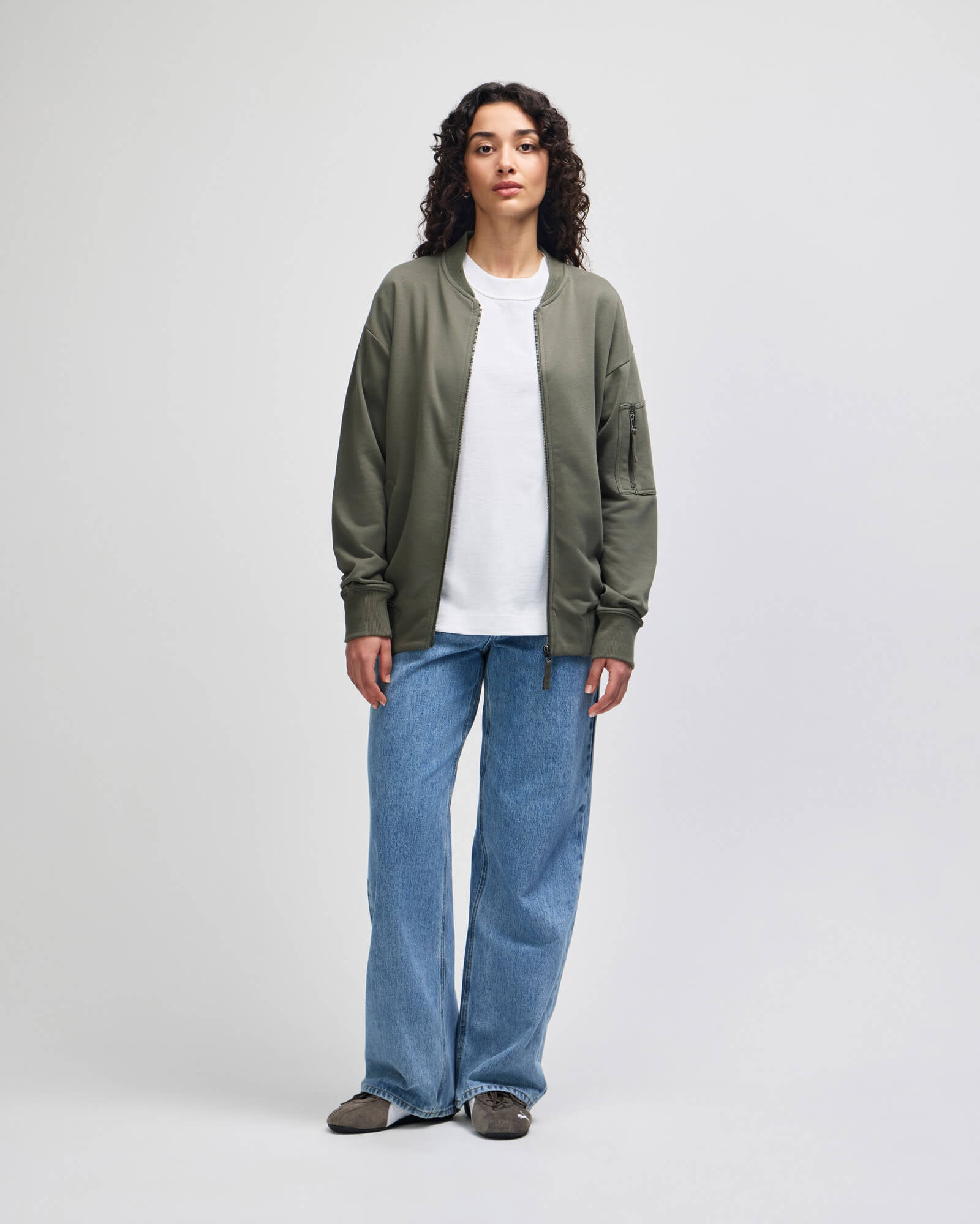 A model stands against a light gray studio background wearing an olive green bomber jacket over a white crewneck tshirt, paired with wide leg blue jeans and dark brown shoes-lieutenant