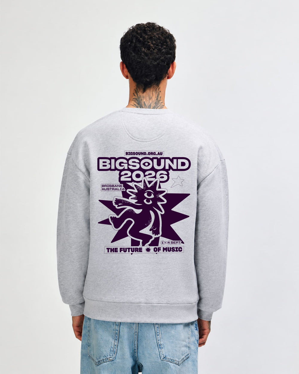 next image of BIGSOUND Crewneck Sweatshirt : A man viewed from behind wearing a light gray crewneck sweatshirt with a large dark purple graphic on the back reading 'BIGSOUND 2025' and 'THE FUTURE OF MUSIC', paired with light blue jeans against a plain white studio background -Heather Gray