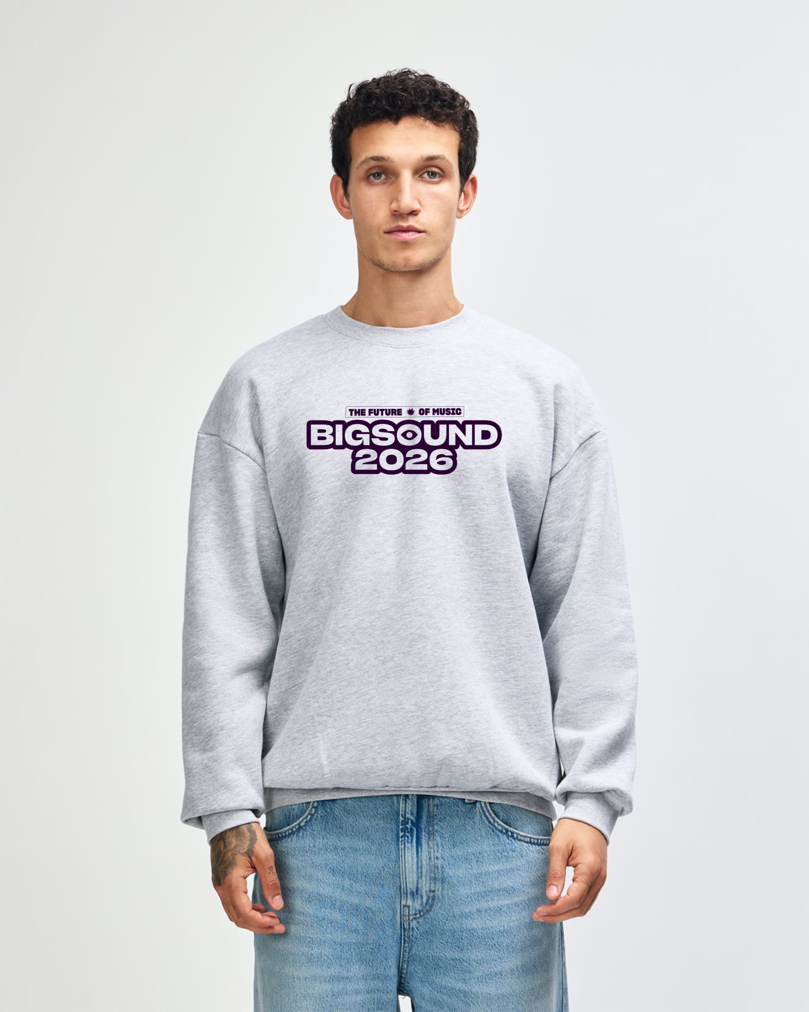A man wearing a light gray crewneck sweatshirt with a 'BIGSOUND 2026' graphic on the chest, paired with blue jeans, standing against a light gray studio background -Heather Gray