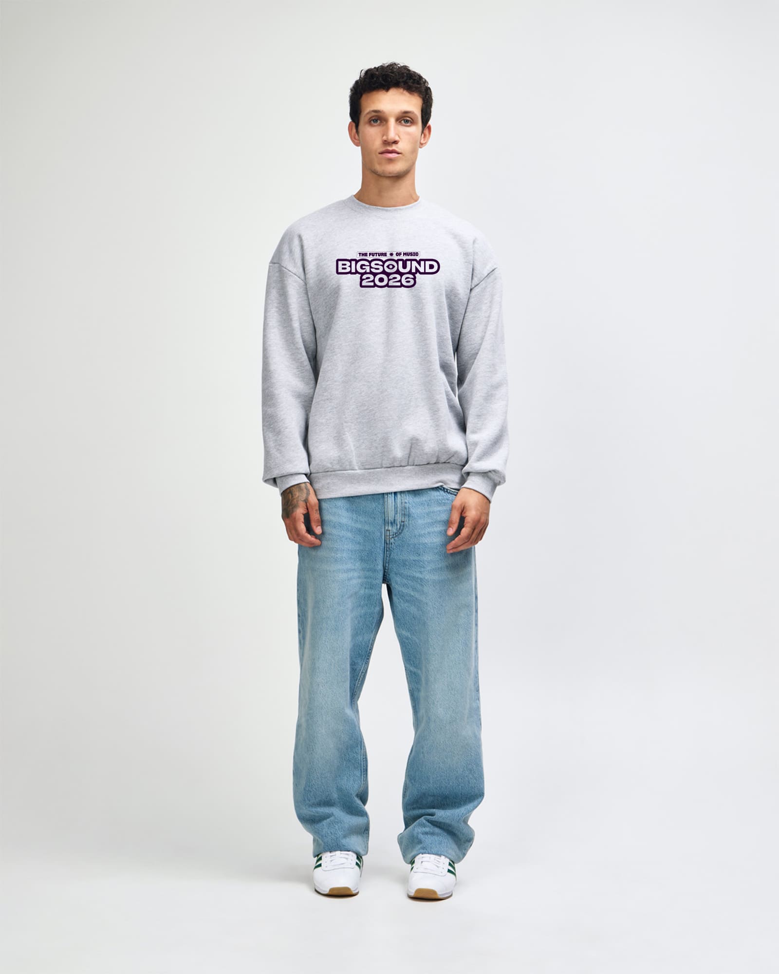A man stands against a light gray studio background wearing a light gray sweatshirt with a purple and black 'BIGGROUND 2026' graphic on the chest, light blue jeans, and white sneakers -Heather Gray